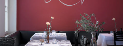 The dining room of Pomodoro. The wall is a deep red color with white cursive writing. The tables are donned in white linens with a single pink rose as the centerpiece of each table. The tables are lined with black chairs.