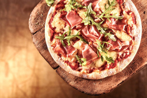a detail shot of a pizza pie. The pizza rests on a wood plank. The pizza is topped with red sauce, fresh melted mozzarella, prosciutto and arugula.