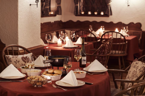 interior shot of a rustic dining room. The walls are white with little candles lining the wooden window sills. The round tables are donned in burgundy linens and wooden country chairs with white patterned cushions circle the tables. Each table has wine classes and elegantly folded napkins at each seat. On the table in the foreground is a bottle of white wine, a fondue pot of melted cheese with potatoes, bread and other dishes to dip in the cheese.