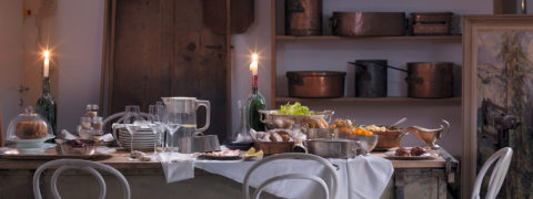 a private dinner table set up in the wine cellar. On the wall are copper pots lined on the wooden shelves as decoration. The long table is full with bowls of bread, grapes and cheeses. There is a bottle of wine in the middle of the table next to a tall candlestick. Plates and wine glasses are stacked on one end of the table across from the food. White chairs line the table.