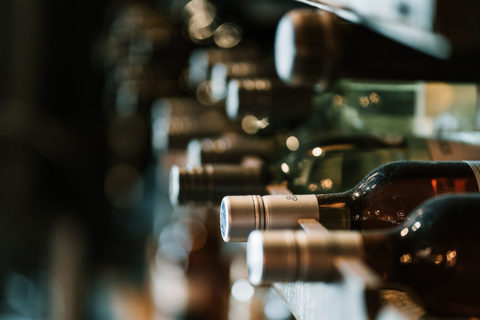 detail shot of wine lined up along a wine cellar shelf. The bottles are different colors with different tops and labels.