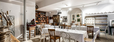 An interior shot of a white dining room. There is decorative silverware lining the shelved on the wall. There are three long tables donned in white linens. Lining the tables are white and brown alternating chairs.