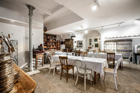 An interior shot of a white dining room. There is decorative silverware lining the shelved on the wall. There are three long tables donned in white linens. Lining the tables are white and brown alternating chairs.