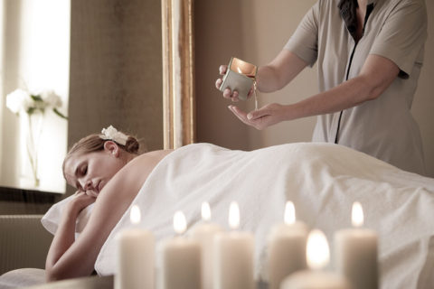 a woman lies on her stomach on a massage table. Her face is tilted toward the camera, her eyes are closed and she looks relaxed. Another woman in a gray uniform stands over her and pours a liquid on her hands to massage into the woman's back