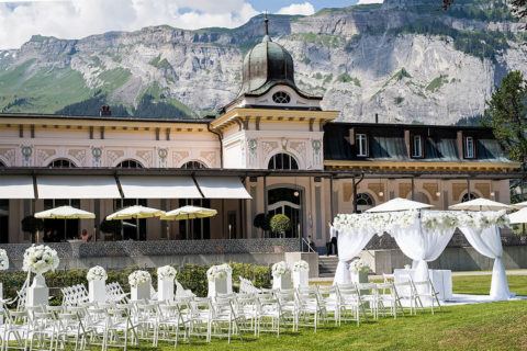 landscape shot of the Waldhaus Flims spa building set up for an outside wedding. There are rows of white chairs leading to a podium surrounded by white drapery and flowers. In the distance are mountains and a blue sky.