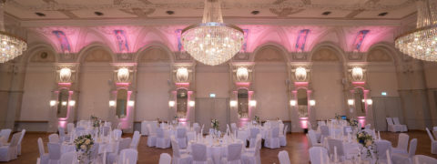 An interior shot of the Belle Epoque Hall decorated for a reception. There are many round tables donned in white linens, elegantly set with large white and green floral centerpieces. There are white chairs circling the tables. Hanging from the ceiling are three large crystal chandeliers.