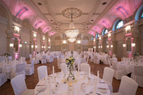 An interior shot of the Belle Epoque Hall decorated for a reception. There are many round tables donned in white linens, elegantly set with large white and green floral centerpieces. There are white chairs circling the tables. Hanging from the ceiling are three large crystal chandeliers.