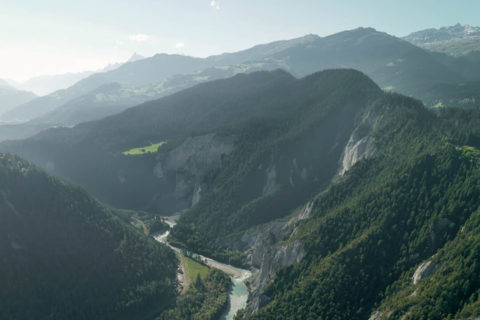 aerial view of the swiss alps during the summer. The mountains are lush green from the evergreen tress and there is a bright blue river running through the valley between the mountains. The sun is shining over the ridges.