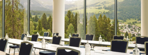 a boardroom with rows of long gray tables and blue seats. At each seat there is a glass bottle of water, a cup a pad of paper and a pen. From the floor to ceiling windows there is a view of evergreen trees, a swiss village and the mountains in the distance.