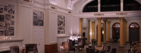 An interior shot of the Belle Epoque Pavilion. At the far end of the room are golden pillars. There are circular brown tables around the room with brown leather chairs with pillows circling each table. There is a fireplace on the left wall. Along the wall are vintage photographs of scenes from the resort.