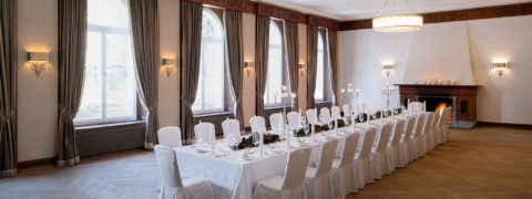 An interior shot of the salon set up for a dinner reception. There is a long table donned with white linens in the center of the room. The table is elegantly set with red and green arrangements running down the middle of the table. There are candlesticks placed in the center of the table as well. At the far end of the room a fireplace is lit.