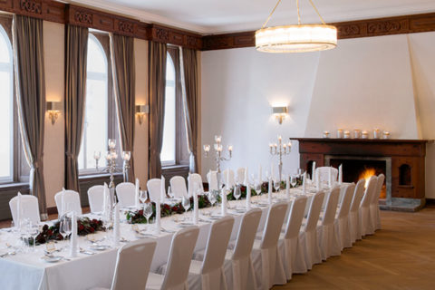 An interior shot of the salon set up for a dinner reception. There is a long table donned with white linens in the center of the room. The table is elegantly set with red and green arrangements running down the middle of the table. There are candlesticks placed in the center of the table as well. At the far end of the room a fireplace is lit.