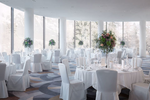 A room set up for a wedding reception. There are many round tables donned in white linens with large flower centerpieces. There are white chairs around every table. The floor to ceiling windows allow a view of evergreen trees and mountain tops covered in snow.