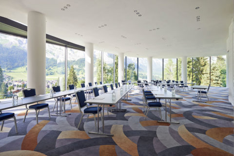 A large boardroom with multiple white long tables in rows. There are black chairs lining both sides of all tables. There is a pad of paper and a pen at each seat. There are green and clear glass bottles of water in the middle of the tables. There is a retro carpet with orange and blues in the pattern. There are floor to ceiling windows that give a view out onto the grounds of the resort and the evergreen forest and mountains in the distance.
