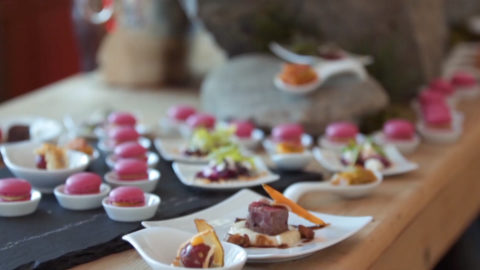 a wooden table is set up with lots of mini dishes. There are meat appetizers on white square plates, pink mini macaroon placed in small white bowls and sauces poured into oversize soup spoons.