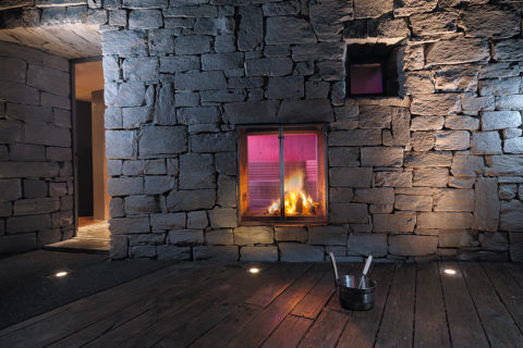 a blazing fire sits in a glass fireplace built into the gray stone wall of a sauna.