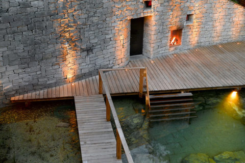 aerial view of the wooden walkway that leads out of the spa and into the pond. The pond is a light green color and there is a fire lit in a fireplace next to the building.