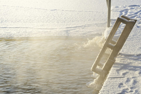 A close up shot of a ladder covered in snow leading out of a steaming hot spring. The water is a dark blue and there is golden tinted steam rising from the surface. The sun reflects off the steam and the snow surrounding the pool