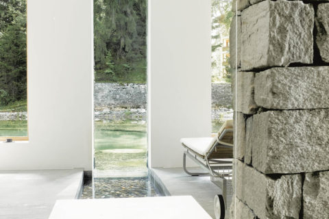 a view from the indoor pool looking out onto the outdoor pool. There is a white lounge chair looking out onto the view.