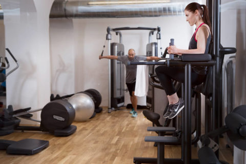 A woman in leggings and a tank top does leg raises on a black machine in the foreground. In the background a man in a gray tshirt strengthens his arms on another black machine. There are ellipticals in the corner of the room