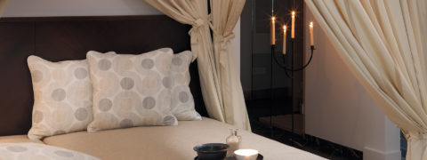 an interior shot of a private spa. There is a beige cushioned bed with gray and beige pillows. The bed is framed by cream colored drapes. There is a platter of two bowls and a glass contained on the bed. There are candle sticks lit in the corner for ambiance.