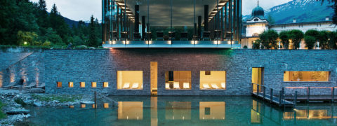 an exterior shot of the resort's spa and outdoor pool. The building is modern and layered. The pool is natural, blending seamlessly into the surrounding nature. There are golden lights coming from the windows of the spa and it is reflecting off of the pool. In the distance are the evergreen trees and tops of the swiss alps.