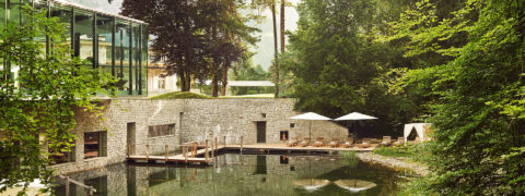 Landscape shot of the resort's pool. The pool is surrounded by lush greenery. The pool leads into the spa and there is a wooden bridge that leads guests inside to the outside and vice versa. Lining the pool are brown lounge chairs and white umbrellas. In the background there is a view of the swiss alps