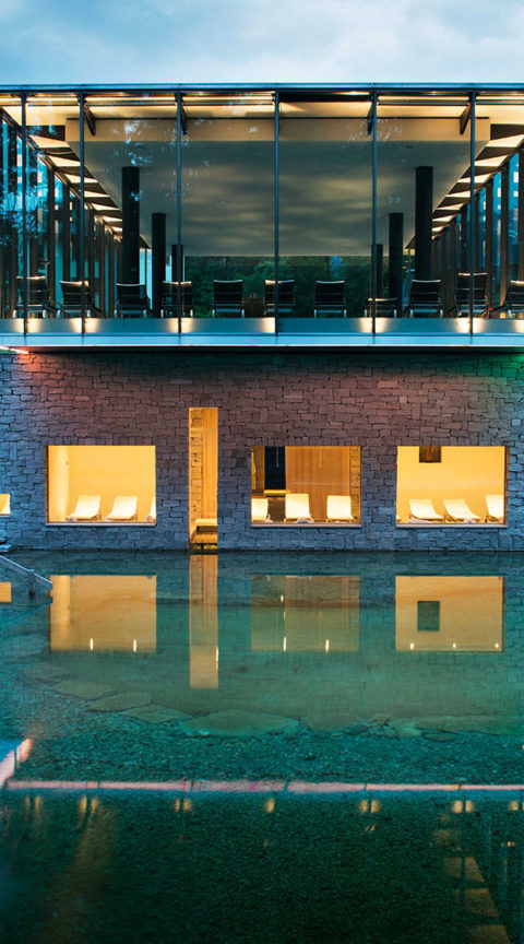 an exterior shot of the resort's spa and outdoor pool. The building is modern and layered. The pool is natural, blending seamlessly into the surrounding nature. There are golden lights coming from the windows of the spa and it is reflecting off of the pool. In the distance are the evergreen trees and tops of the swiss alps.