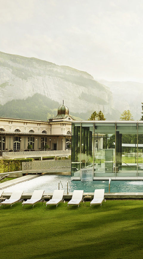 exterior shot of the waldhaus flims spa building and pool. The building is white with decorative fixtures at the top. The pool is indoor and outdoor. The indoor portion of the pool is encased in a glass fixture. The outside portion of the pool is lined with white lounge chairs. In the distance are evergreen trees and the swiss alps.