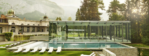 exterior shot of the waldhaus flims spa building and pool. The building is white with decorative fixtures at the top. The pool is indoor and outdoor. The indoor portion of the pool is encased in a glass fixture. The outside portion of the pool is lined with white lounge chairs. In the distance are evergreen trees and the swiss alps.