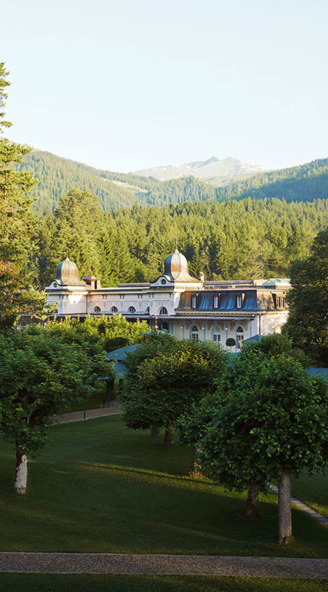 a landscape shot of the Waldhaus Flims resort building and the surrounding grounds. The building is white and is contrasted by the bright green of the evergreen trees that surround it.