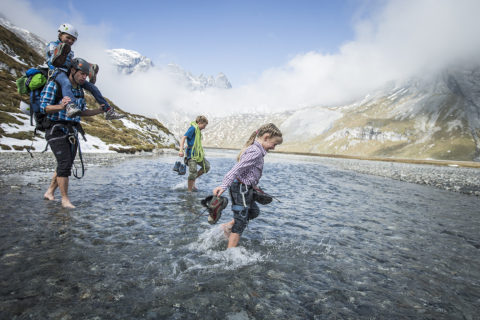 dad with 3 kids walking barefoot through stream of water in between mountains