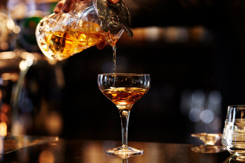 a bartender pours a caramel colored drink into a martini glass