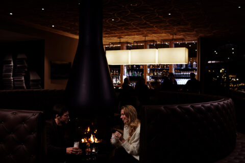 a couple sitting in the bar by the fireplace enjoying drinks and lively conversation, in the background there is the main bar fill with people having drinks and fun