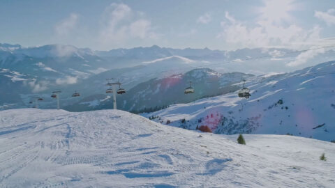 drone panning view of the ski lifts, man in red ski outfit skiing and makes a stop to look around the beautiful mountain top view, he then continue to ski down the mountain. Now night time, we see the couple in front of the outdoor fireplace having hot tea with sparkling lights around the resort and the ski mountain in the background